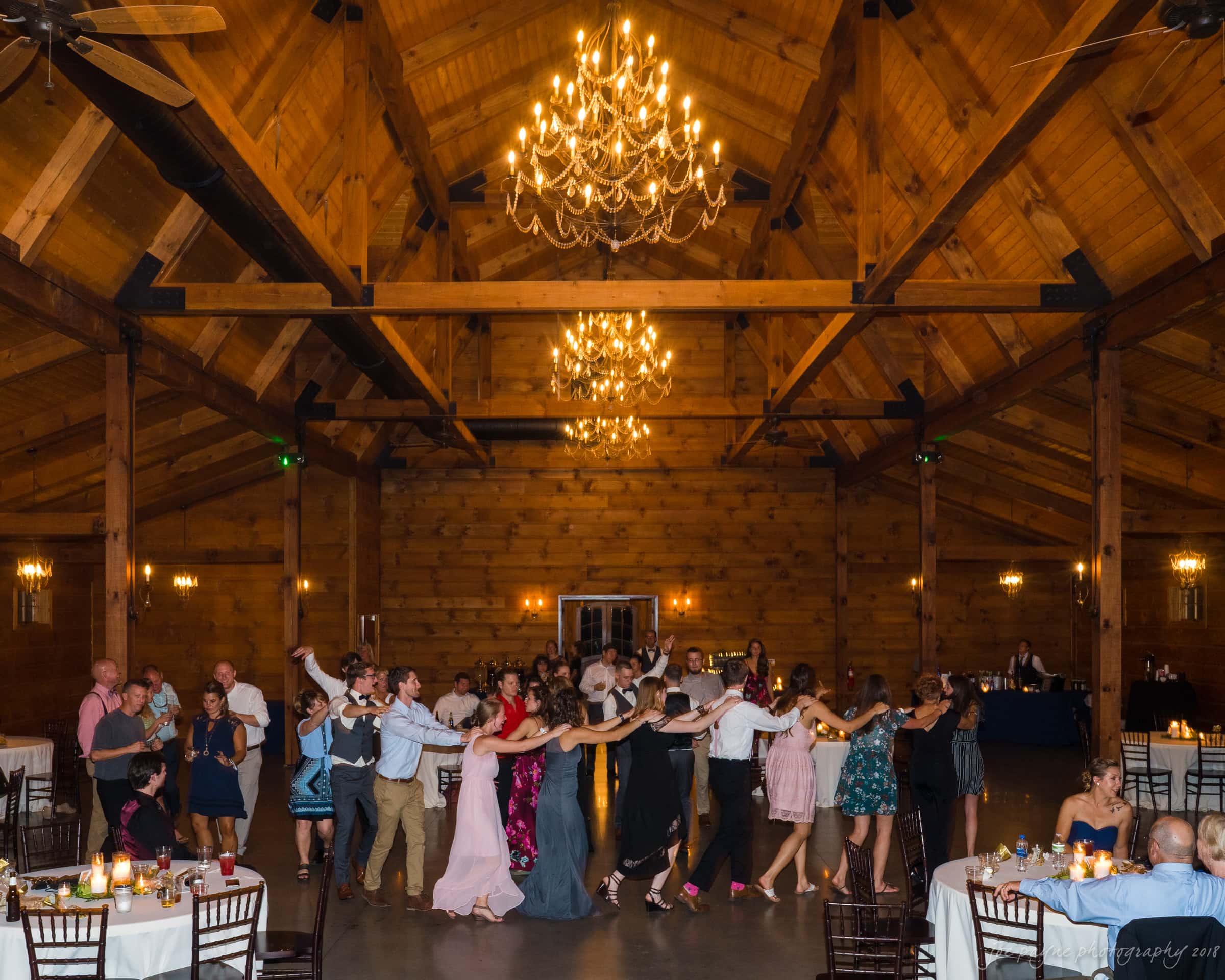 pavilion at carriage farm wedding - ashley & devon A lively group of people forms a conga line in a spacious wooden hall with chandeliers overhead. Tables with candles are set around the room, and guests dance joyfully, dressed in formal attire, creating a festive atmosphere.