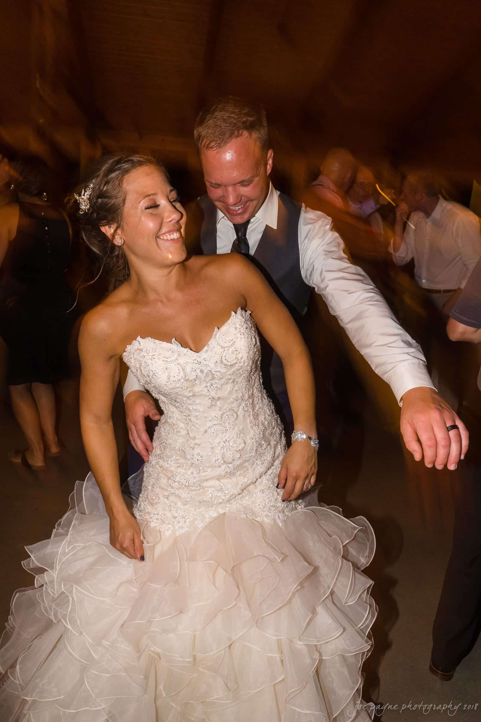 pavilion at carriage farm wedding - ashley & devon A smiling bride in a lace wedding dress dances joyfully with a groom in a vest and tie. The motion blur suggests theyre spinning, surrounded by other guests in a warm, festive setting.