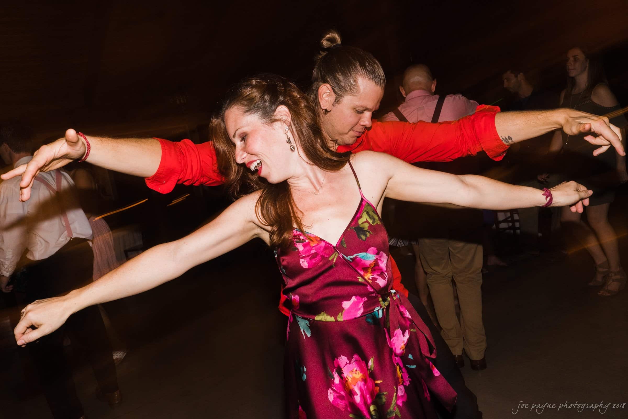 pavilion at carriage farm wedding - ashley & devon A woman in a floral dress and a man in a red shirt are dancing joyfully with their arms outstretched on a dimly lit dance floor. Other people are visible in the background, also engaged in dancing and socializing.