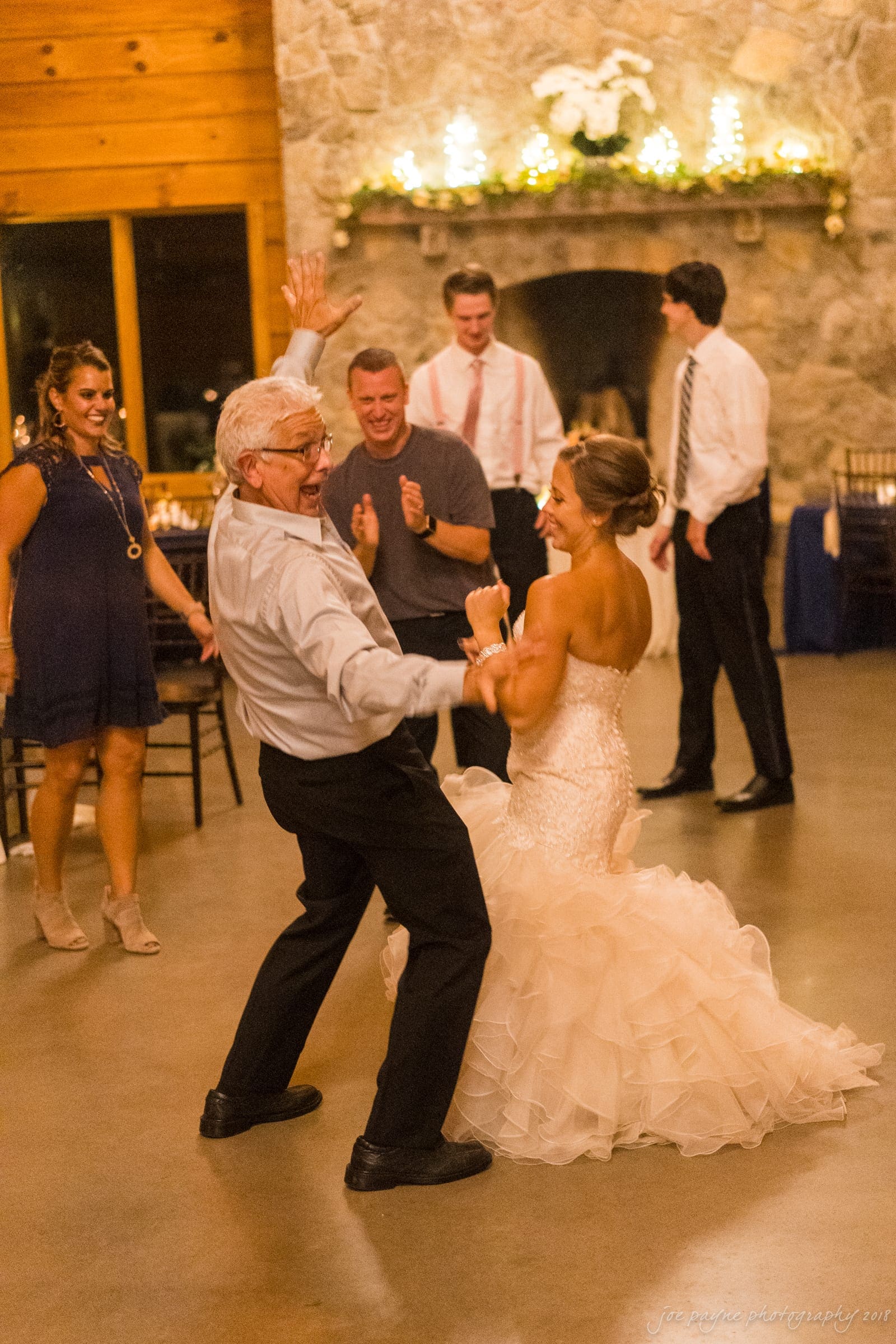 pavilion at carriage farm wedding - ashley & devon A bride in a strapless gown dances joyfully with an older man in a shirt and tie. Several guests in dresses and suits cheer them on. The background features a stone wall and soft, warm lighting, creating a festive atmosphere.