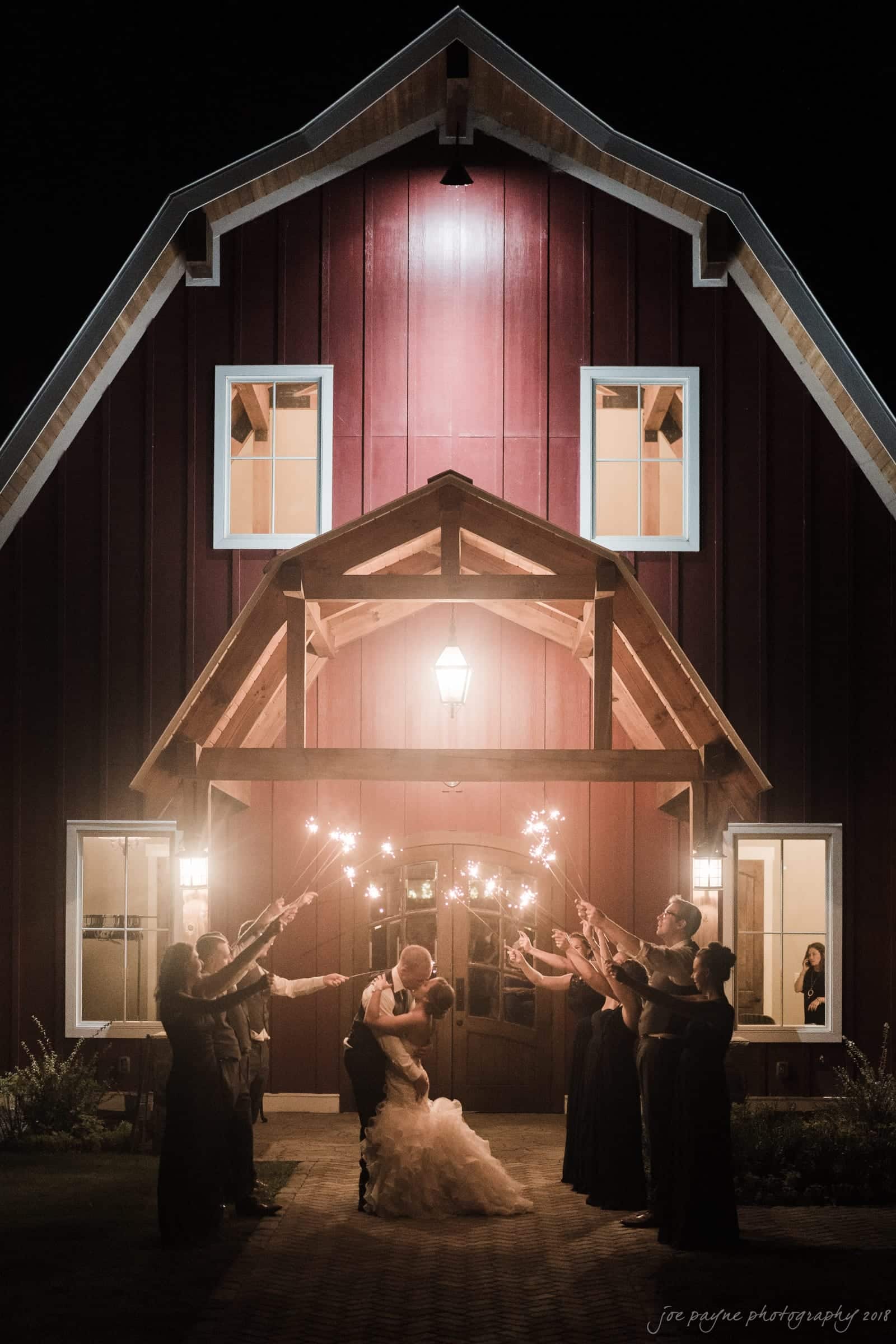 pavilion at carriage farm wedding - ashley & devon A bride and groom share a kiss under a wooden porch lit by lanterns. They are surrounded by a wedding party holding sparklers. The scene is set in front of a red barn with warm glowing windows, creating a romantic and festive atmosphere.