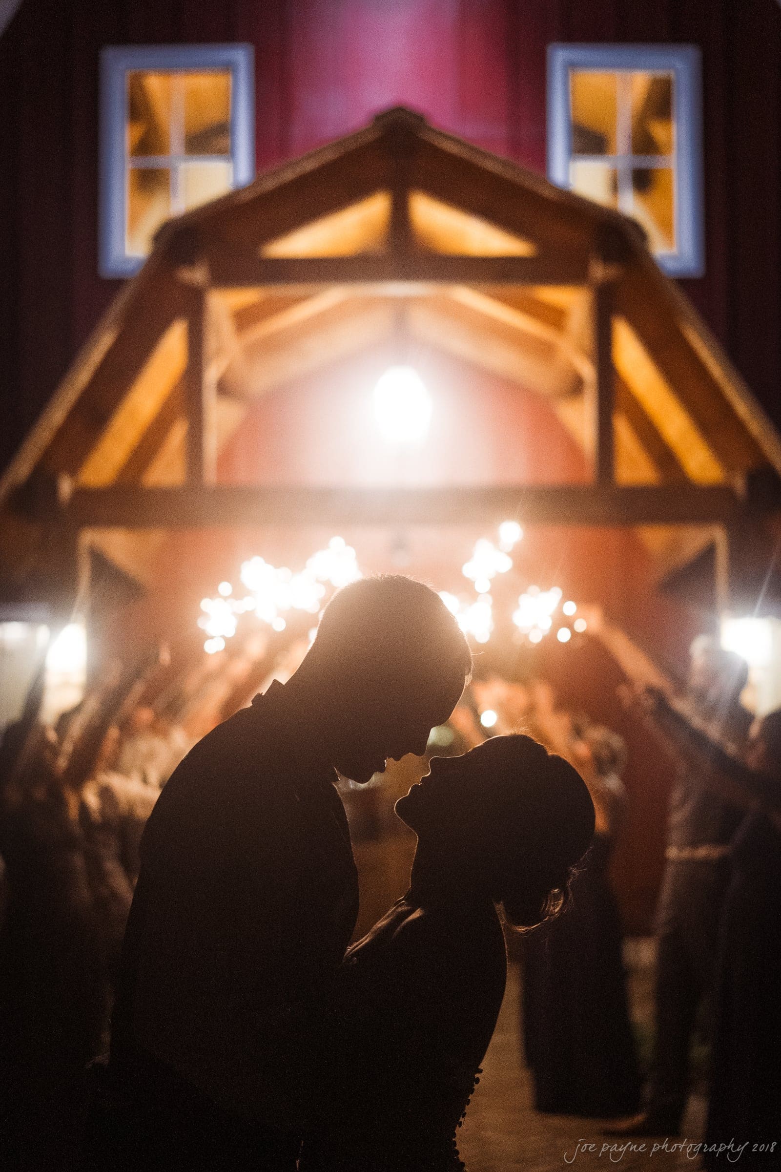 pavilion at carriage farm wedding - ashley & devon A silhouetted couple embraces in front of a warmly lit barn entrance. Sparkers illuminate their outline, while a group of people holding sparklers forms an arch around them, creating a romantic and festive atmosphere.