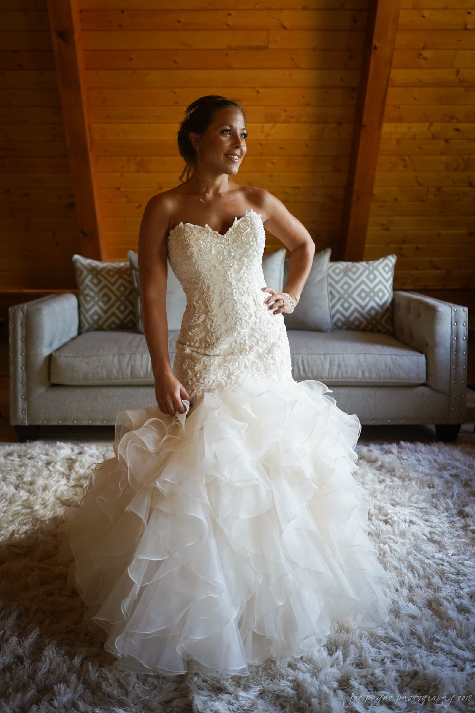 pavilion at carriage farm wedding - ashley & devon A woman in a white strapless wedding dress with a ruffled skirt stands on a plush carpet in a cozy room. Wooden walls and a gray couch with patterned pillows are in the background. The woman is looking to the side and smiling.