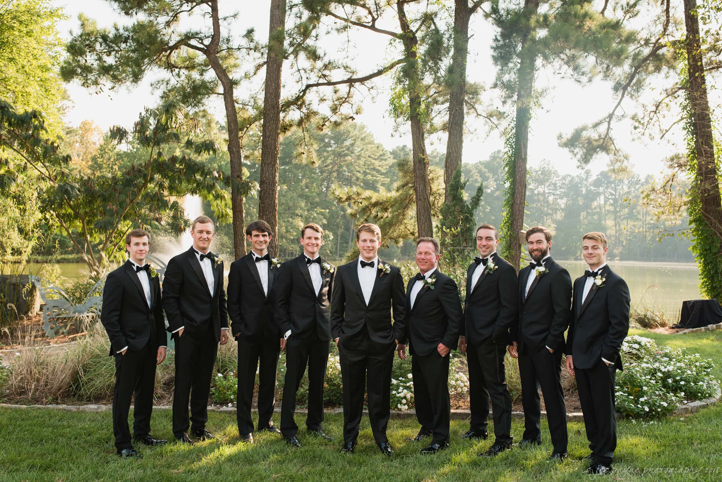 A group of nine men in tuxedos stand on a lawn, with trees and a lake in the background. The setting appears formal, possibly for a wedding or special event. The men are smiling and pose together under dappled sunlight.