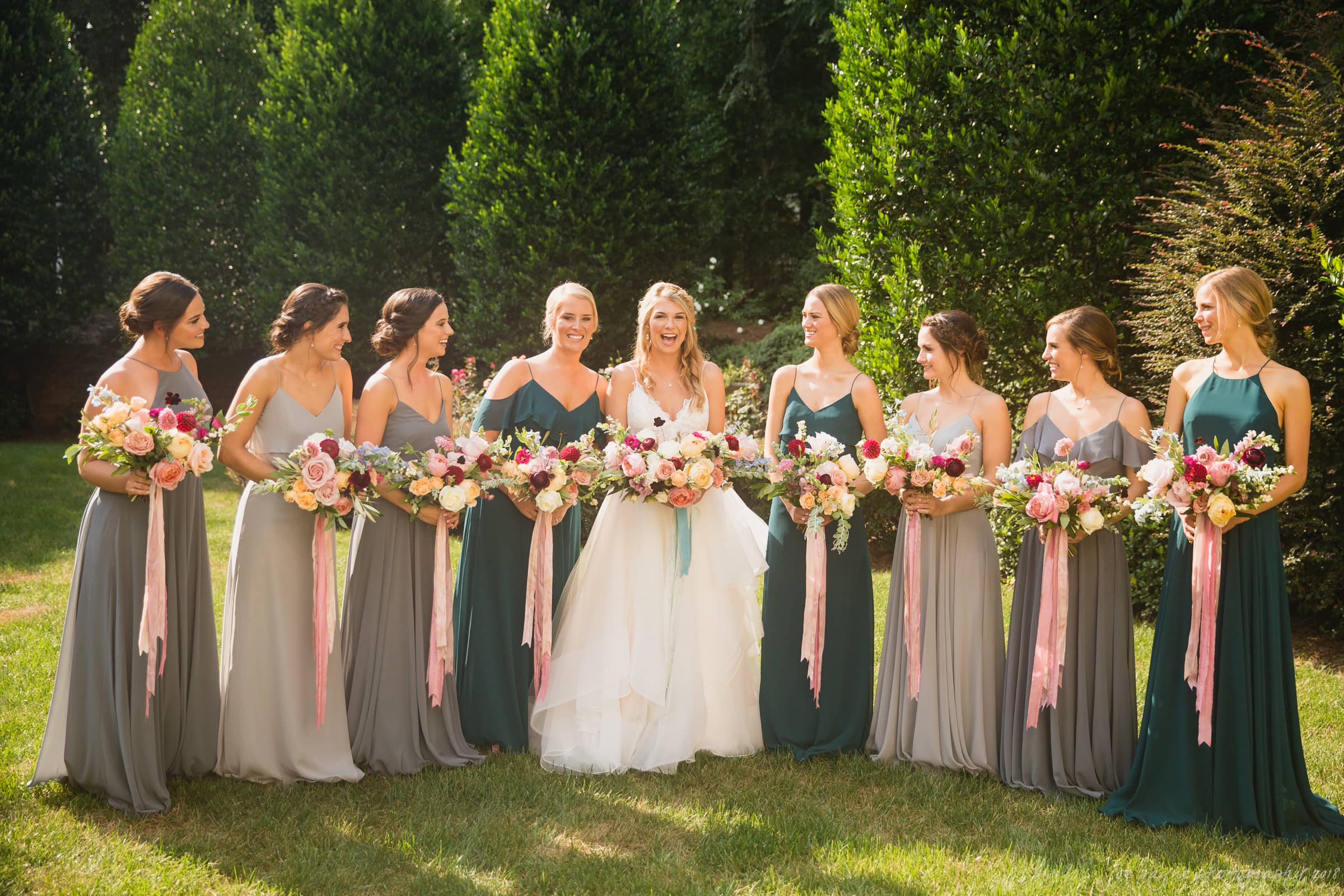 A bride stands smiling with eight bridesmaids outdoors in a garden. They are holding colorful bouquets and wearing long dresses in shades of gray and teal. They are surrounded by lush green trees.