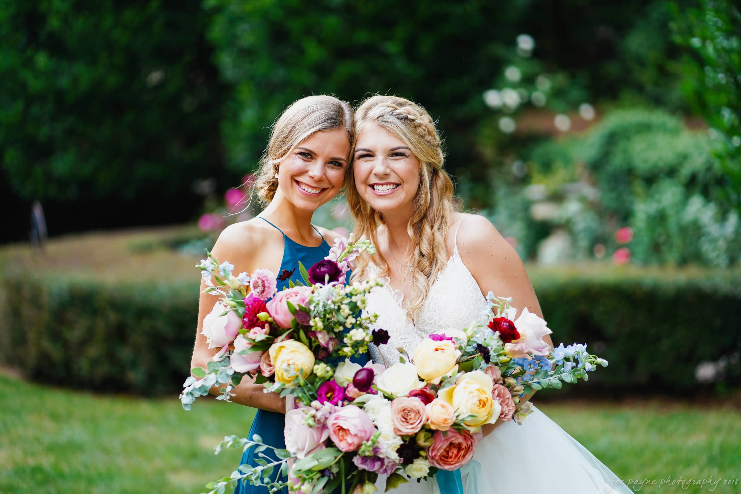 Two smiling women stand together outdoors, each holding a vibrant bouquet of flowers. The woman on the left wears a teal dress, while the woman on the right is in a white dress. A lush, green garden serves as the backdrop.