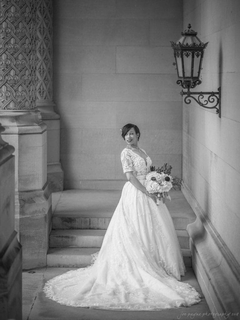 biltmore wedding photographer - anneké & andrew A bride in an elegant white lace gown stands on stone steps, holding a bouquet. She is smiling and looking to her left. The setting is ornate with intricate details on the walls and a decorative lantern above.