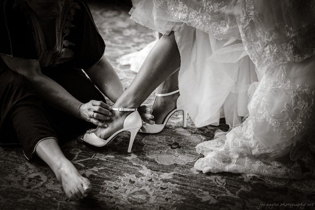 biltmore wedding photographer - anneké & andrew A black-and-white photo of a bride sitting as another person helps fasten the ankle strap of her high-heeled shoes. The brides lace wedding dress is visible, and both are seated on a patterned rug.