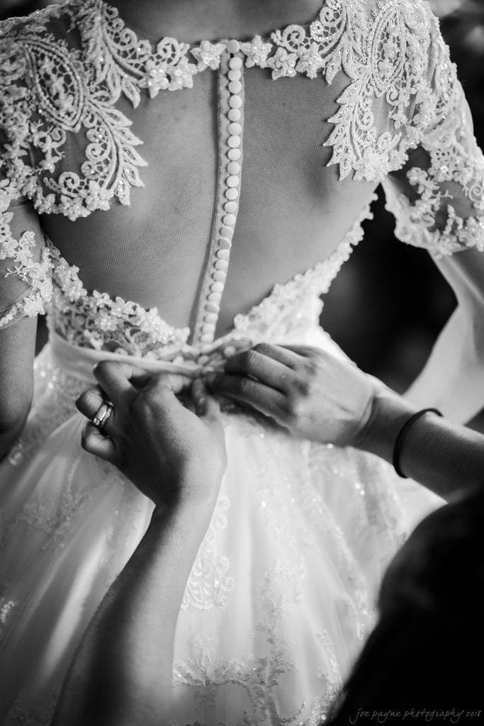 biltmore wedding photographer - anneké & andrew A black-and-white photo shows someone fastening the back buttons of an intricate lace wedding gown. The dress features a detailed lace design and an open back with a long row of buttons.