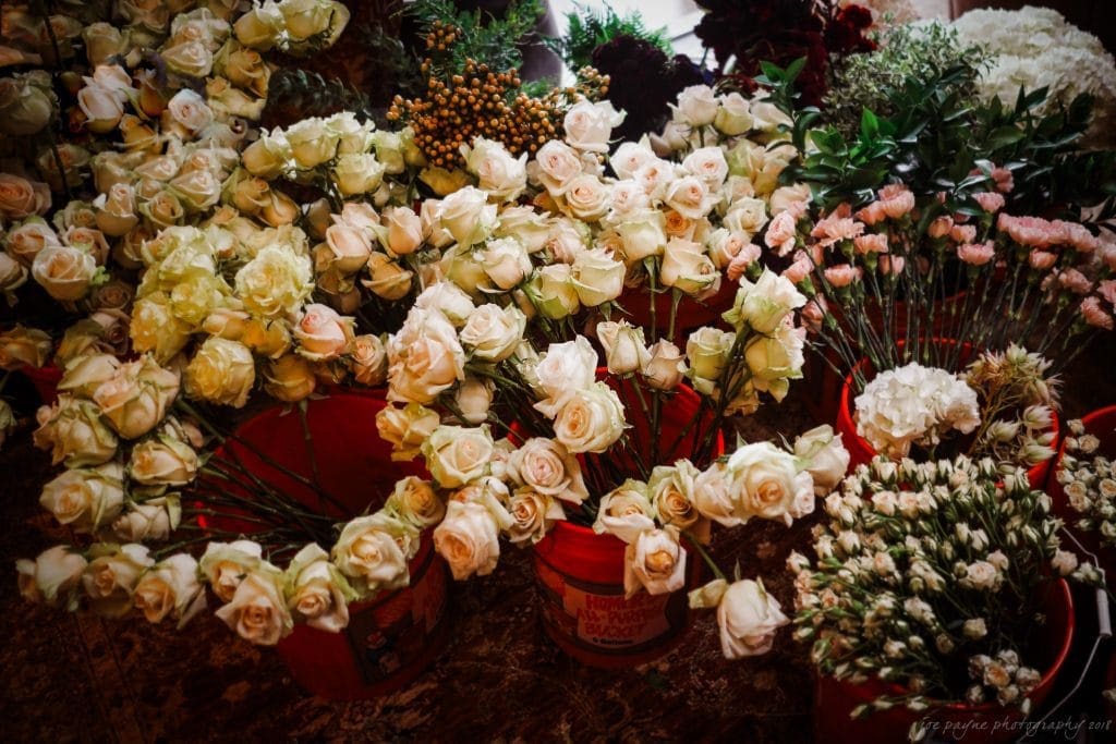 biltmore wedding photographer - anneké & andrew Buckets filled with a variety of flowers, including cream and pale pink roses, are arranged on a surface. The background shows additional bunches of greenery and small blooms, creating a lush, vibrant floral display.