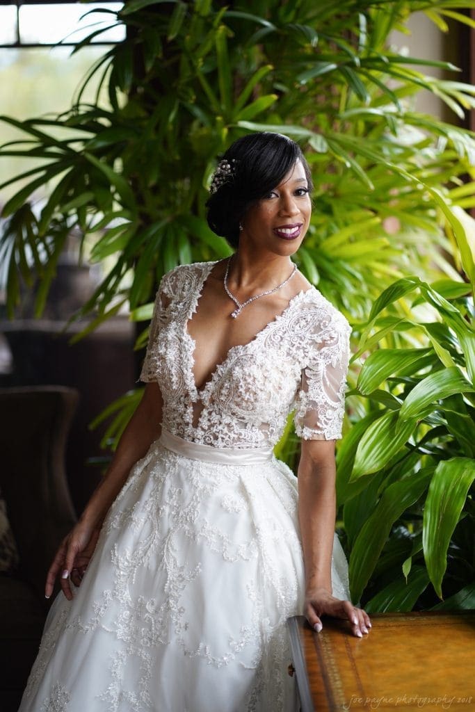 biltmore wedding photographer - anneké & andrew Bride in a lace wedding dress stands in front of lush green plants. She has short, styled hair adorned with a decorative accessory and is smiling gently, leaning slightly against a wooden surface. The lighting is soft and natural.