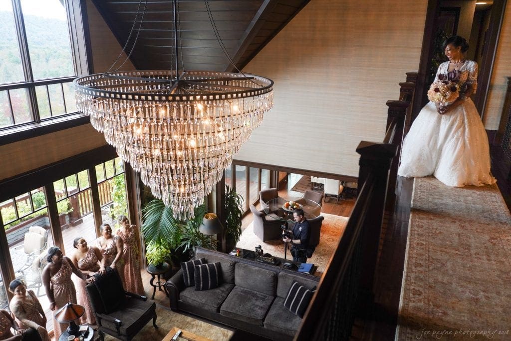biltmore wedding photographer - anneké & andrew A bride in a white gown stands on a balcony holding a bouquet, overlooking a room with a grand chandelier, where bridesmaids in matching dresses gather. Large windows reveal a scenic outdoor view.