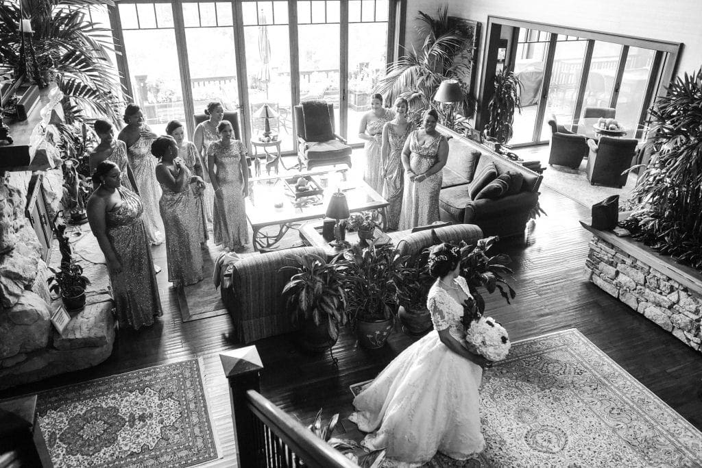 biltmore wedding photographer - anneké & andrew A bride in a flowing gown and veil holds a bouquet as she walks through a living room. Bridesmaids, dressed in matching gowns, watch from across the room. Large windows and greenery enhance the elegant setting.