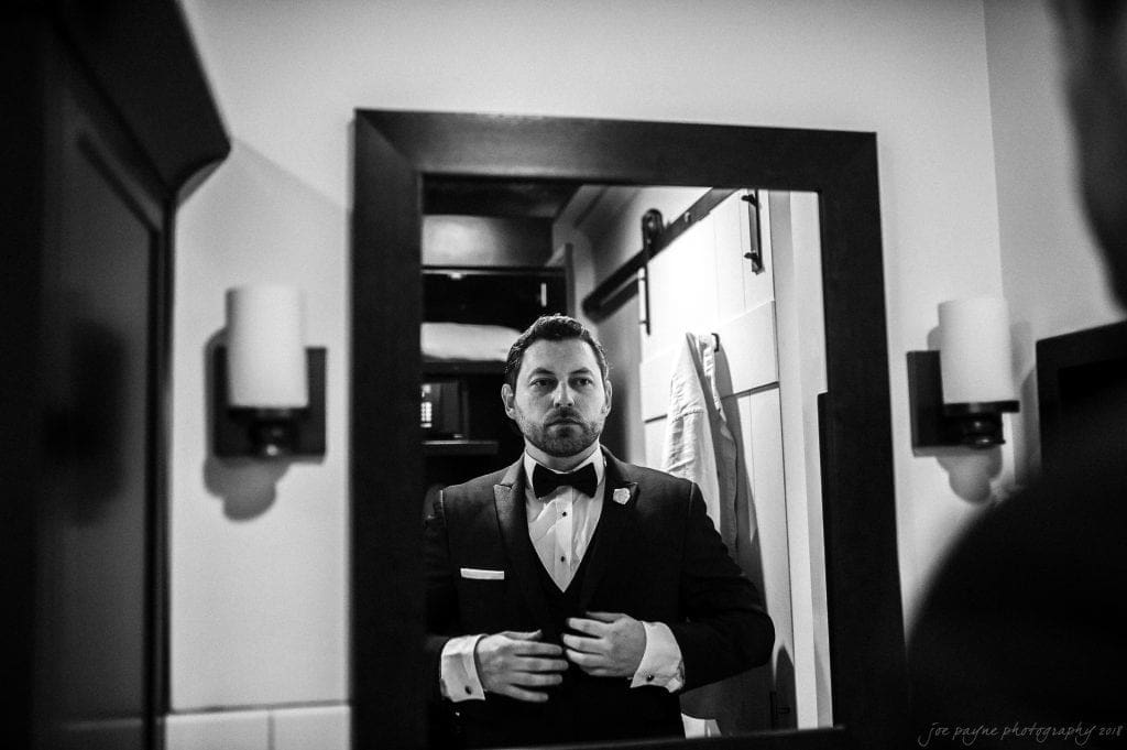 biltmore wedding photographer - anneké & andrew A man in a tuxedo adjusts his bow tie while looking in a mirror. The room is softly lit, and a suit jacket is hanging on the wall in the background. The image is in black and white.