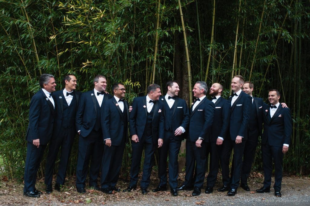 biltmore wedding photographer - anneké & andrew A group of eleven men in formal black suits and bow ties stand together outdoors in front of tall bamboo plants. They are smiling and interacting with each other.