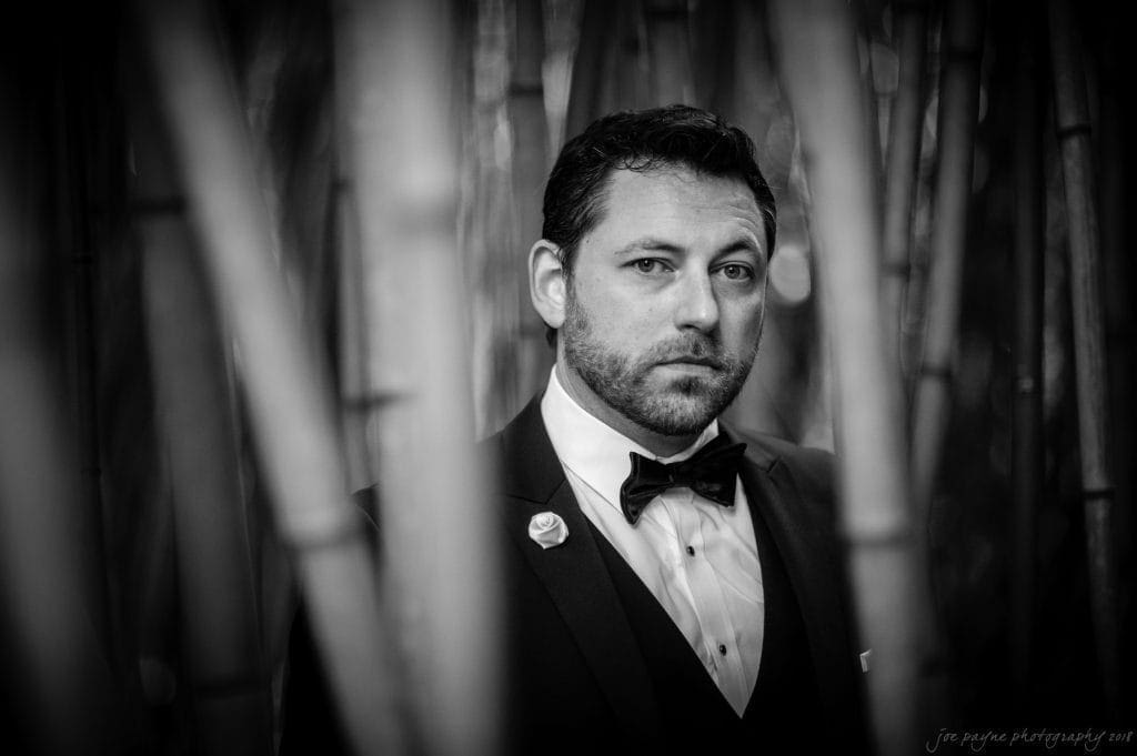biltmore wedding photographer - anneké & andrew A man in a tuxedo and bow tie stands among tall bamboo stalks. The image is in black and white, giving it a classic, timeless feel. He gazes directly at the camera with a thoughtful expression.