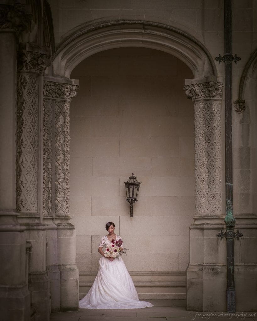 biltmore wedding photographer - anneké & andrew A bride in a white gown stands under an ornate archway, holding a bouquet of flowers. The setting features intricate stonework and a vintage lantern, creating a classic and elegant scene.