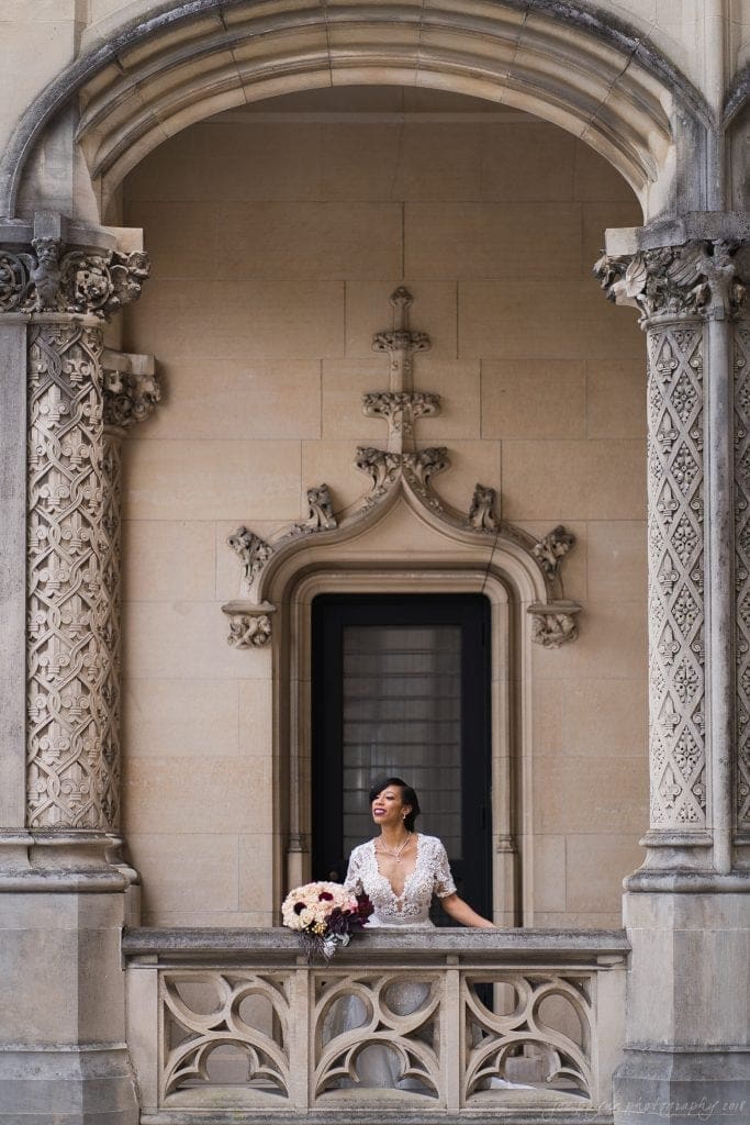 biltmore wedding photographer - anneké & andrew A person in a white dress stands on an ornate balcony, adorned with carved stone columns and arch. They are holding a bouquet of flowers, smiling, and looking upwards. The architectural details are intricate and the setting appears historic.