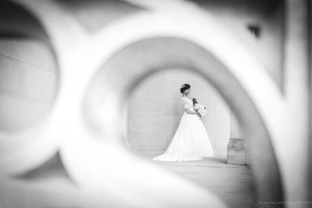 biltmore wedding photographer - anneké & andrew A bride in a flowing white dress holds a bouquet as she stands gracefully within an archway. The image, in black and white, is creatively framed through ornate architectural details, creating an elegant and timeless feel.