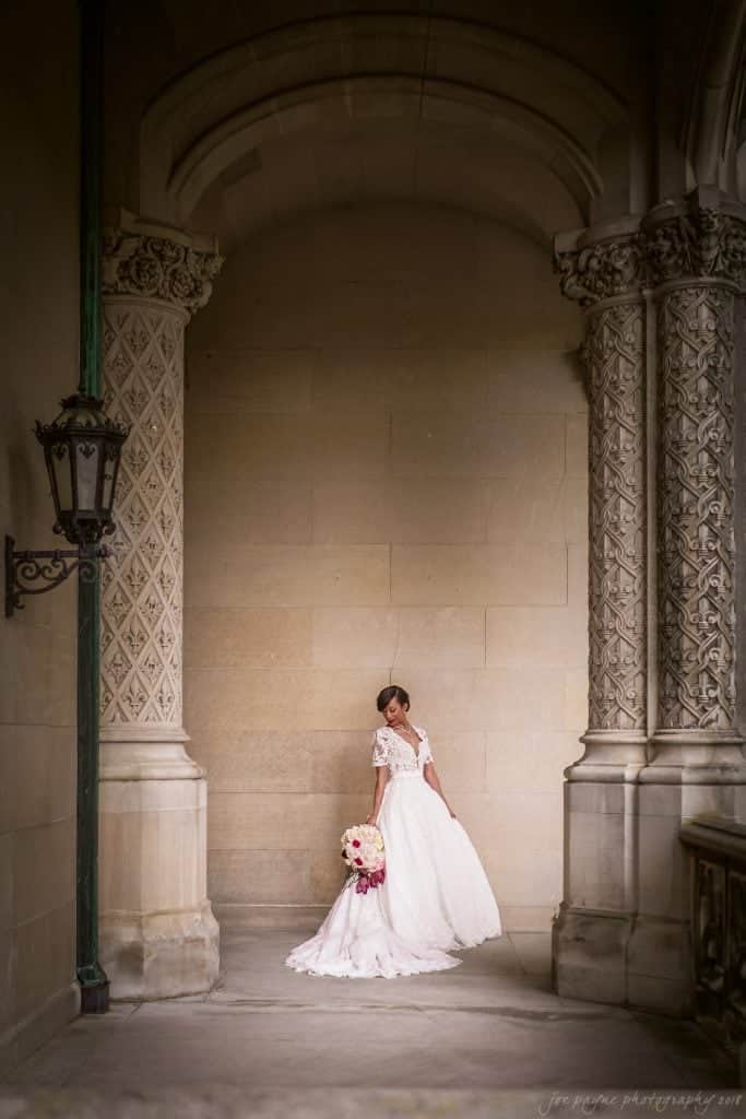biltmore wedding photographer - anneké & andrew A bride in a white dress stands in an ornate, tall archway. She holds a bouquet of pink and white flowers. The walls are decorated with intricate carvings. An elegant lantern is mounted on the left wall.