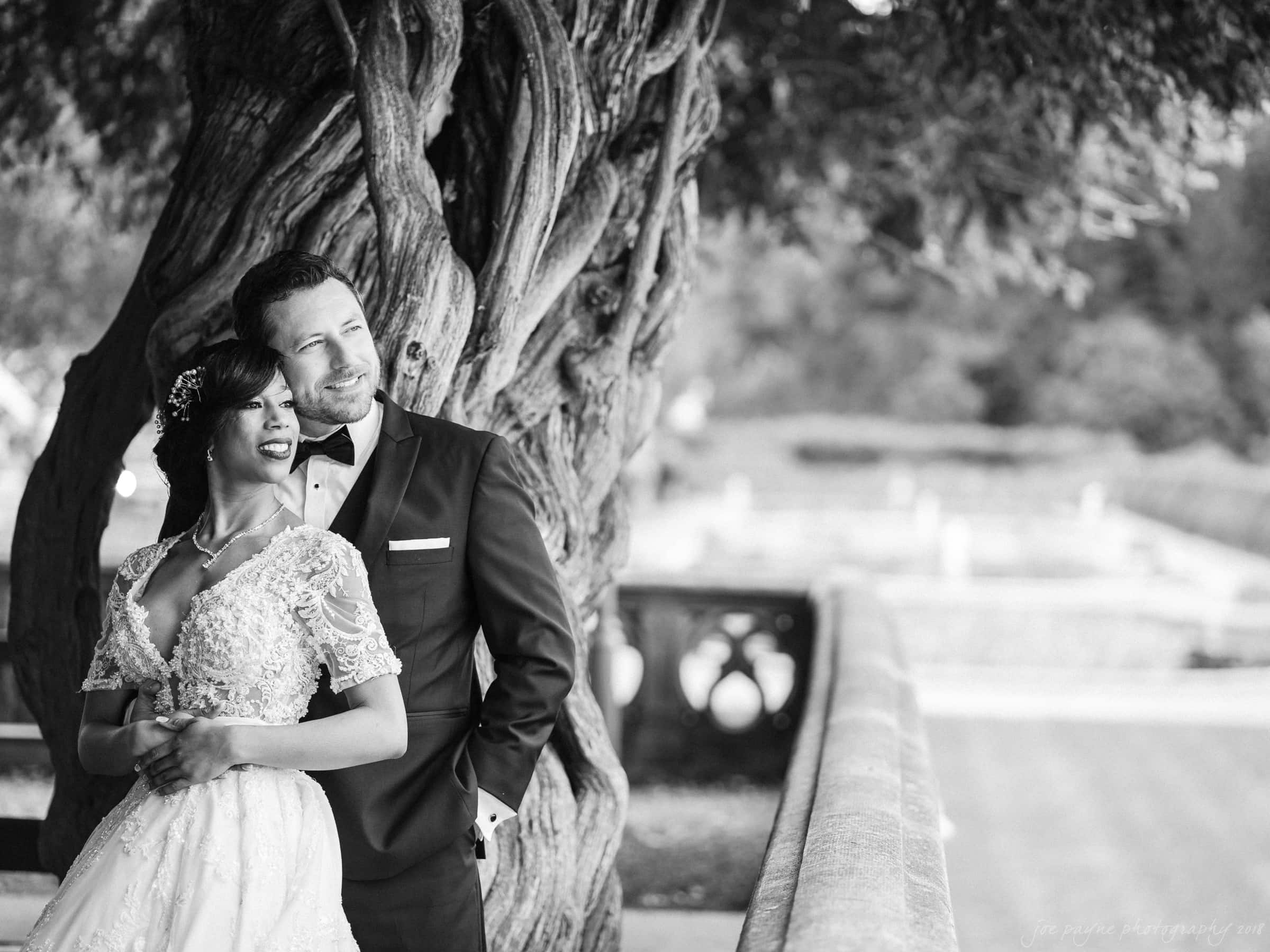 A black and white photo captures a bride and groom embracing, smiling under a large, twisted tree—a quintessential Biltmore wedding scene. The bride dazzles in her lacy gown while the groom looks sharp in his suit, set against a stone path with blurred greenery providing an enchanting backdrop.