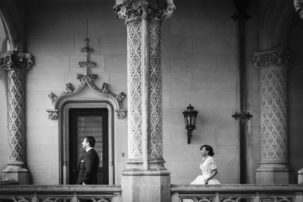 biltmore wedding photographer - anneké & andrew A black-and-white photo of a bride and groom. The groom, wearing a suit, stands in profile, while the bride, in a gown, walks towards him from behind. They are on a decorative balcony with ornate columns and a vintage wall lantern.