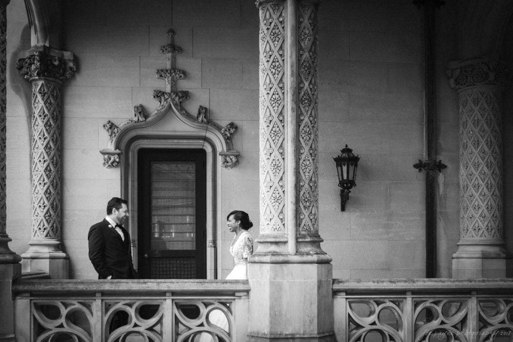 biltmore wedding photographer - anneké & andrew A black-and-white photo of a couple on a balcony. The man is in a suit, and the woman is in a gown. They stand facing each other, surrounded by ornate architectural details, including columns and intricate stonework.