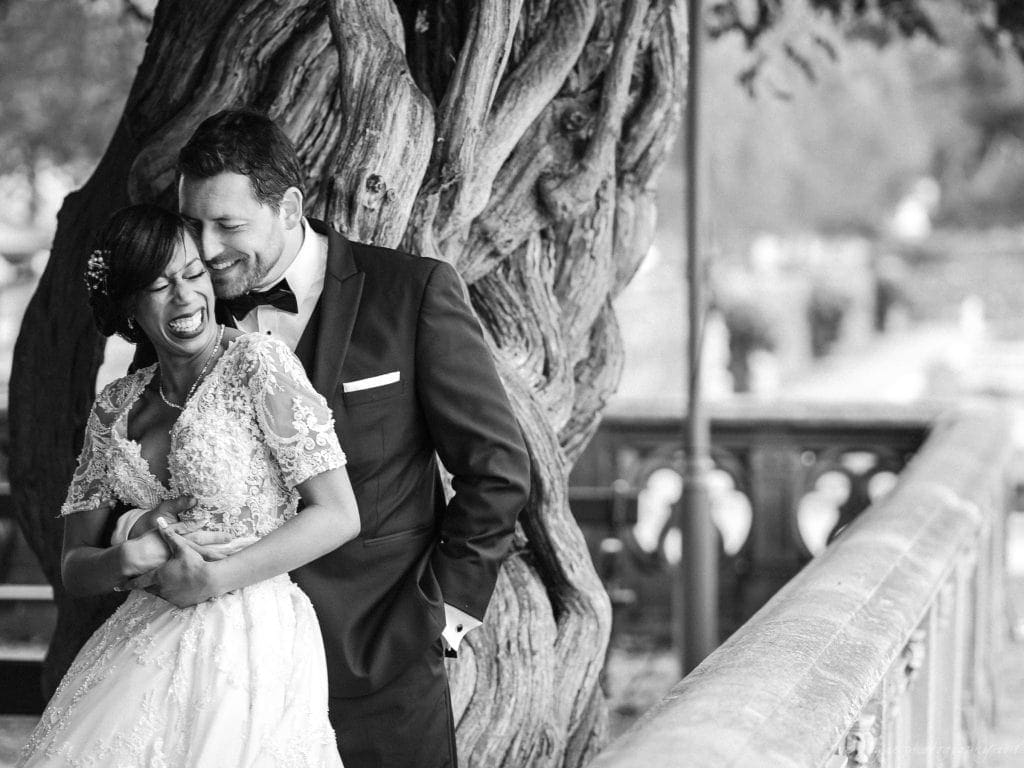 biltmore wedding photographer - anneké & andrew A joyful bride and groom embrace and laugh together beside a gnarled tree. The bride is wearing an elegant lace gown, and the groom is in a dark suit with a bow tie. The scene is serene and romantic, captured in black and white.