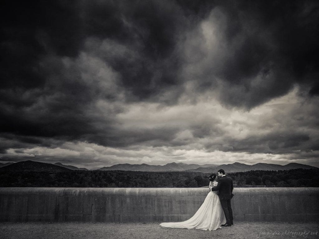 biltmore wedding photographer - anneké & andrew A bride and groom embrace in front of a dramatic, stormy sky. The bride wears a flowing gown, and the groom is in a dark suit. They stand against a backdrop of distant mountains and a vast landscape, creating a romantic and moody scene.