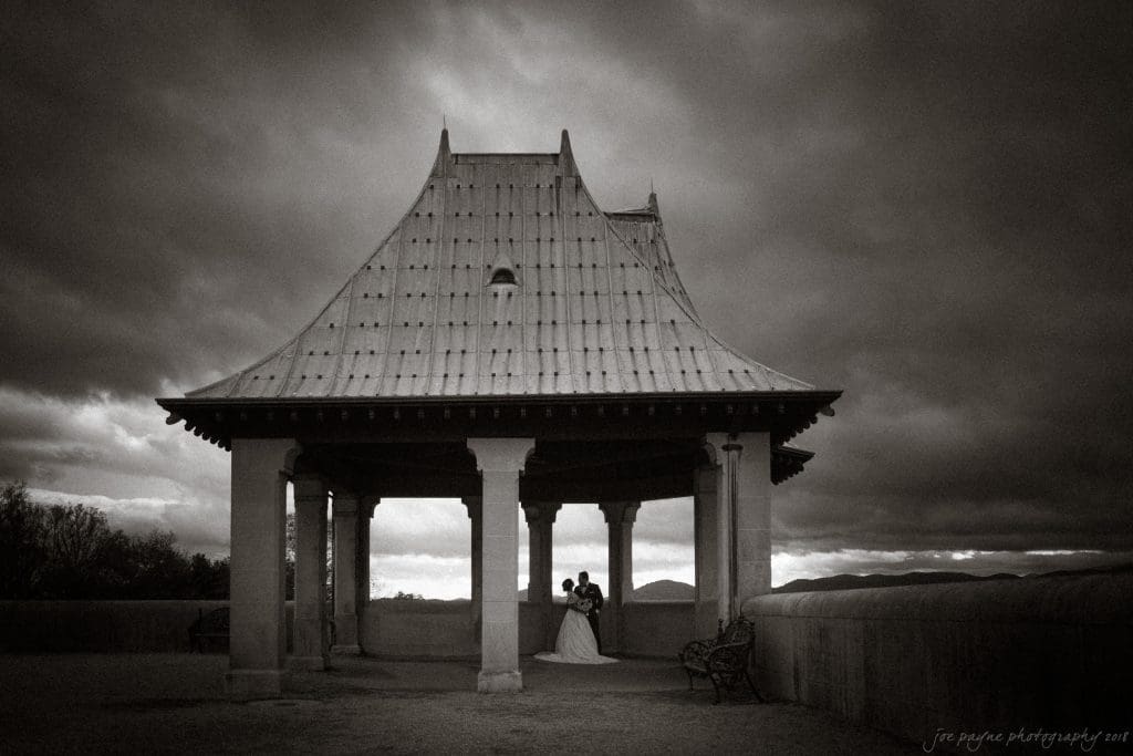 biltmore wedding photographer - anneké & andrew A couple embraces under a large pagoda-style pavilion with a dramatic cloudy sky in the background. The scene is in black and white, creating a moody and romantic atmosphere.