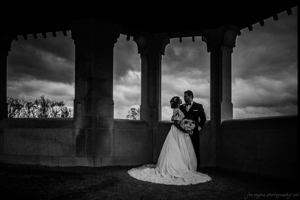 biltmore wedding photographer - anneké & andrew A bride and groom stand under a stone pavilion, framed by arched openings. The bride wears a flowing dress and holds a bouquet, while the groom is in a suit. The sky is overcast, creating a dramatic background.
