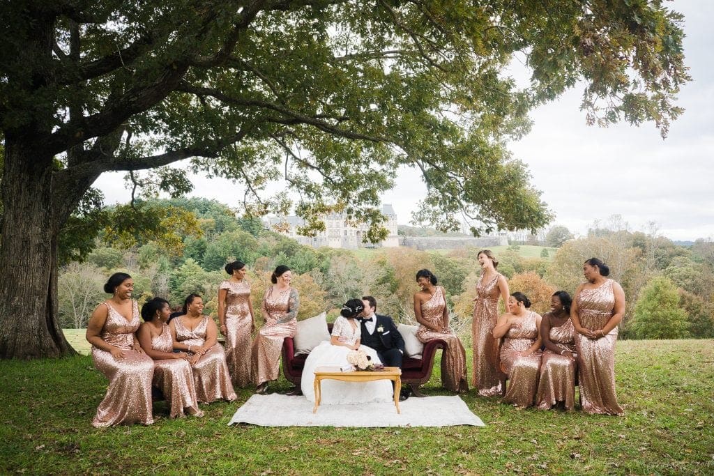 biltmore wedding photographer - anneké & andrew A wedding party poses under a large tree with a bride and groom sitting on a red sofa. The bride wears a white dress, the groom a black tuxedo, and the bridesmaids shimmering bronze dresses. A hilly landscape and a castle are visible in the background.