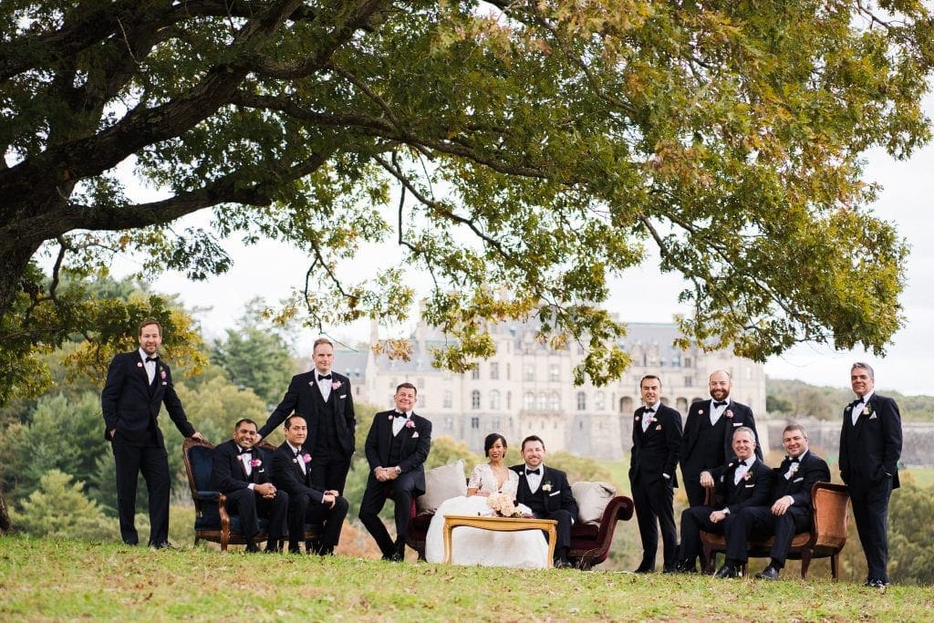 biltmore wedding photographer - anneké & andrew A group of people in formal black attire gather outdoors under a large tree, posing with sofas and a small table. A large building is visible in the background.