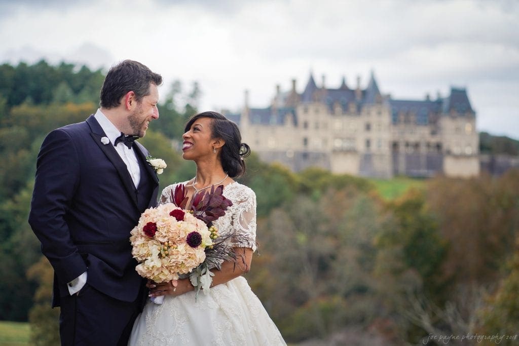 biltmore wedding photographer - anneké & andrew A bride and groom smile at each other while standing outdoors. The bride holds a bouquet of flowers. In the background, there is a large castle-like building surrounded by trees and a cloudy sky.