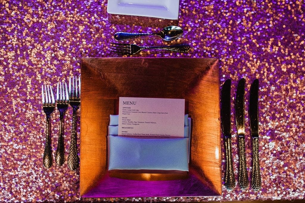 biltmore wedding photographer - anneké & andrew A sparkling gold table setting with a square copper plate, a folded menu, and stylish silverware. The tablecloth glitters in shades of purple and gold. Four forks are on the left and three knives with a spoon on the right.