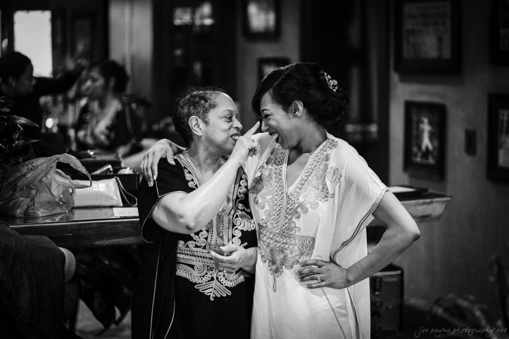 biltmore wedding photographer - anneké & andrew A black and white photo of two women in embroidered dresses sharing a joyful moment, with one playfully touching the others face. Background details include framed pictures on the walls and a table with bags.
