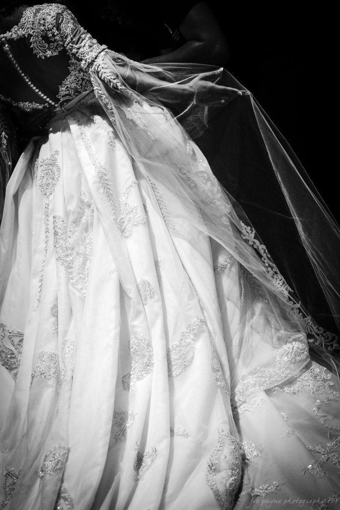 biltmore wedding photographer - anneké & andrew A black and white photo of a bride wearing an ornate wedding gown with intricate lace and embroidery patterns. The veil flows gracefully, creating elegant layers. The background is dark, highlighting the dress details.