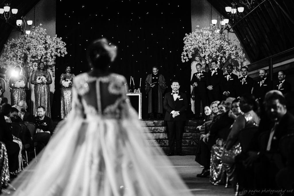 biltmore wedding photographer - anneké & andrew A black-and-white photo of a bride walking down the aisle towards a groom at the altar. The wedding party, in formal attire, stands on either side amid floral decorations. Guests are seated, watching the ceremony. Chandelier lighting sets an elegant scene.
