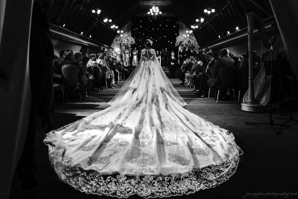 biltmore wedding photographer - anneké & andrew A bride walks down the aisle in an ornate gown with a long, detailed train. The setting is a dimly lit venue with guests seated on either side. The focus is on the brides dress and the dramatic ambiance of the ceremony.