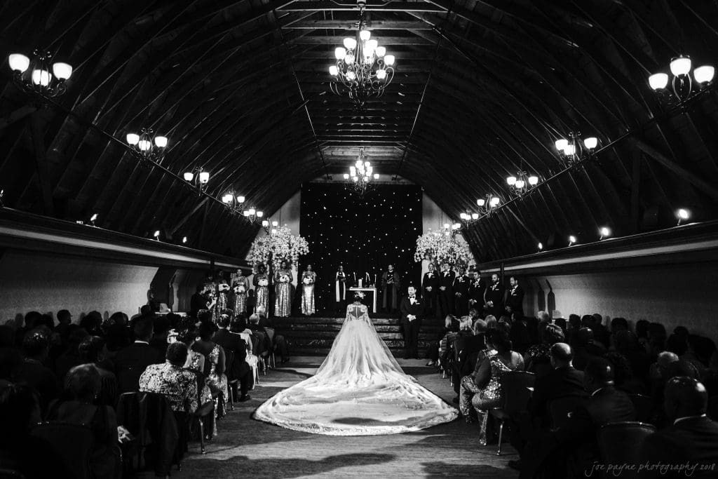 biltmore wedding photographer - anneké & andrew A bride in a long flowing gown walks down the aisle of a grand, dimly lit venue with a high vaulted ceiling and chandeliers. Guests are seated on either side, and an elegantly decorated altar is visible at the front.