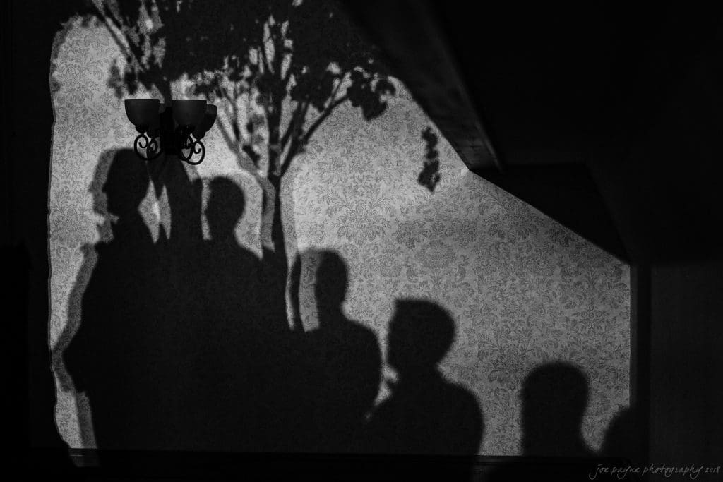 biltmore wedding photographer - anneké & andrew Shadows of several people and a tree are cast on a patterned wall, illuminated by a lamp. The image is in black and white, giving a dramatic and artistic effect.