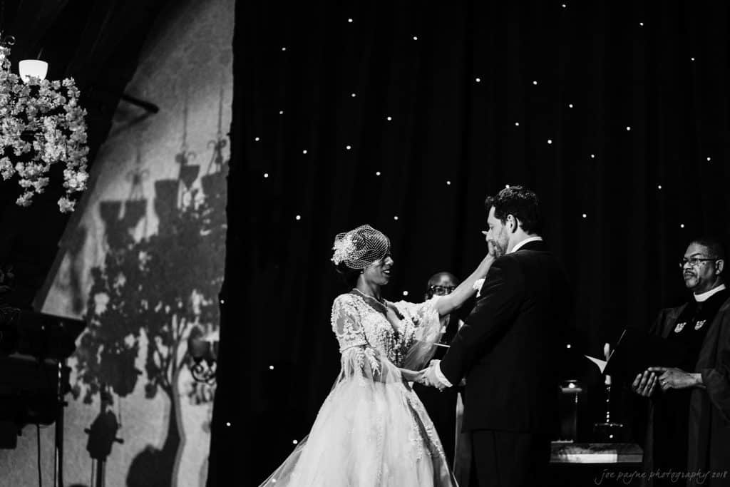 biltmore wedding photographer - anneké & andrew A bride and groom stand facing each other, smiling during their wedding ceremony. The bride wears a lace gown and birdcage veil, while the groom is in a suit. They are holding hands in front of a dark backdrop adorned with small lights.