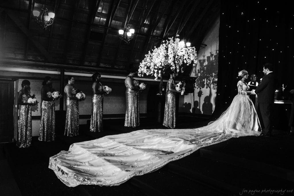 biltmore wedding photographer - anneké & andrew A bride and groom stand together, facing each other under a floral arch. The brides long train is spread out behind her. Six bridesmaids in sequined dresses hold bouquets in a line on the left. The setting is dimly lit, creating a dramatic atmosphere.