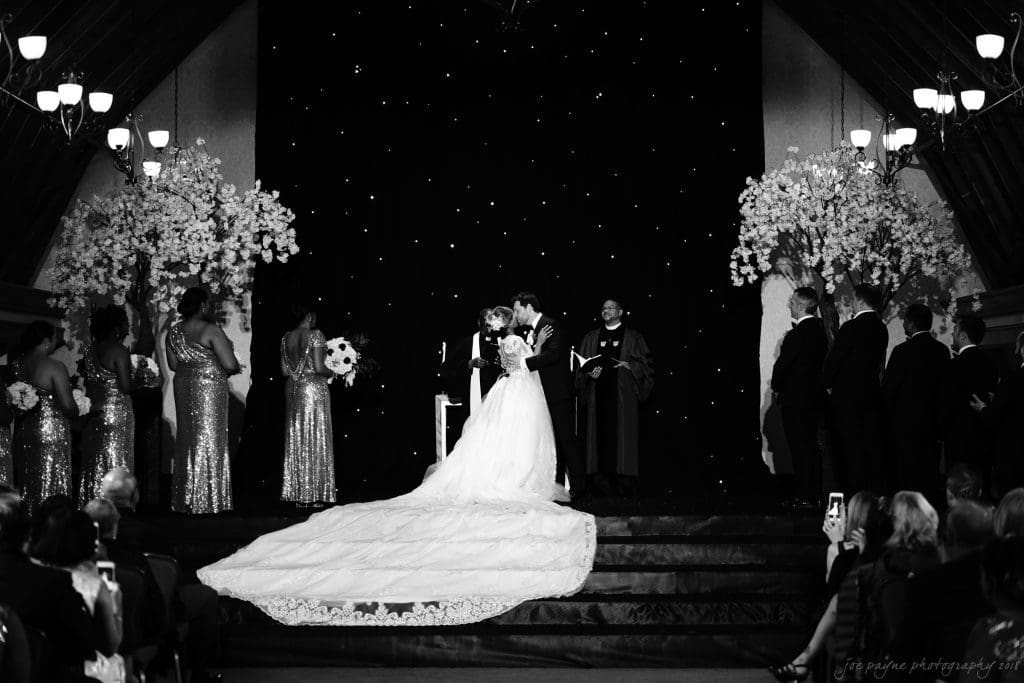 biltmore wedding photographer - anneké & andrew A black-and-white photo of a wedding ceremony. The bride and groom kiss at the altar, surrounded by bridesmaids in sparkly dresses and groomsmen. Large floral arrangements adorn the stage, and guests look on. The brides long train is prominent.