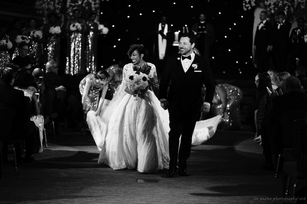 biltmore wedding photographer - anneké & andrew A bride and groom are joyfully walking down the aisle, hand in hand, after their wedding ceremony. The bride wears a flowing gown, and the groom is in a tuxedo. Guests seated on either side are watching and celebrating the couple.