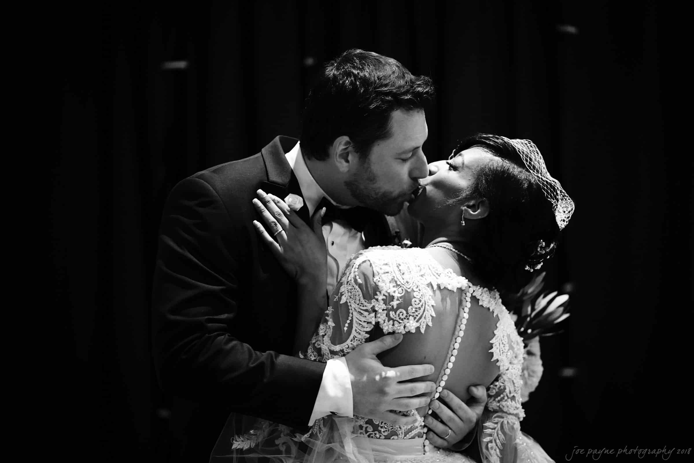 biltmore wedding photographer - anneké & andrew A bride and groom share a kiss on their wedding day. The groom is wearing a tuxedo, and the bride is in a lace wedding dress with a sheer back. They embrace against a dark background.