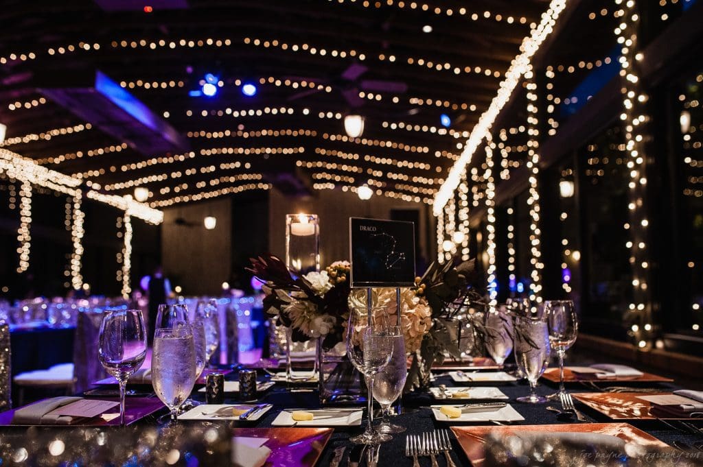 biltmore wedding photographer - anneké & andrew A beautifully decorated reception hall with tables set for an event. Twinkling string lights hang from the ceiling, illuminating elegant place settings with glasses, plates, and floral centerpieces under a dim, cozy ambiance.