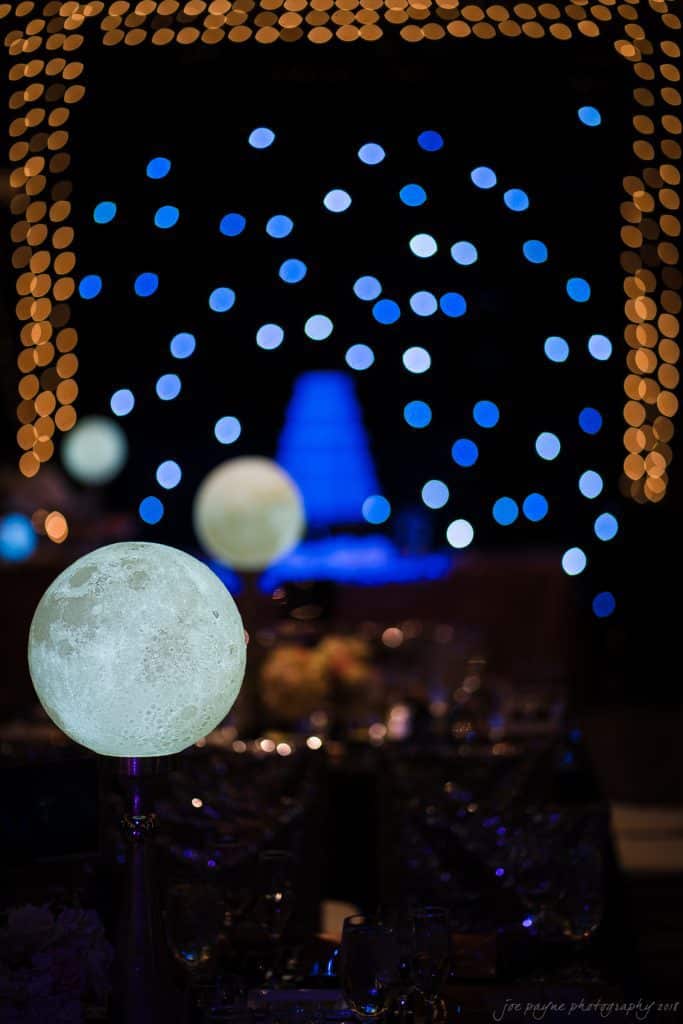 biltmore wedding photographer - anneké & andrew Dimly lit event scene with glowing moon lamps as centerpieces on tables. Background features a soft focus of warm white and cool blue bokeh lights, creating a dreamy atmosphere. Glassware and table settings are visible in the foreground.