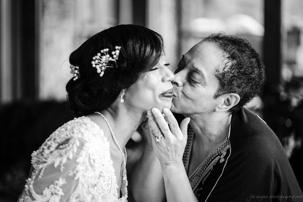 biltmore wedding photographer - anneké & andrew Two people share a joyful moment in a black and white photo. One is wearing a wedding dress with intricate lace and a floral hairpiece, while the other in a dark patterned outfit gently kisses their cheek, both smiling warmly.