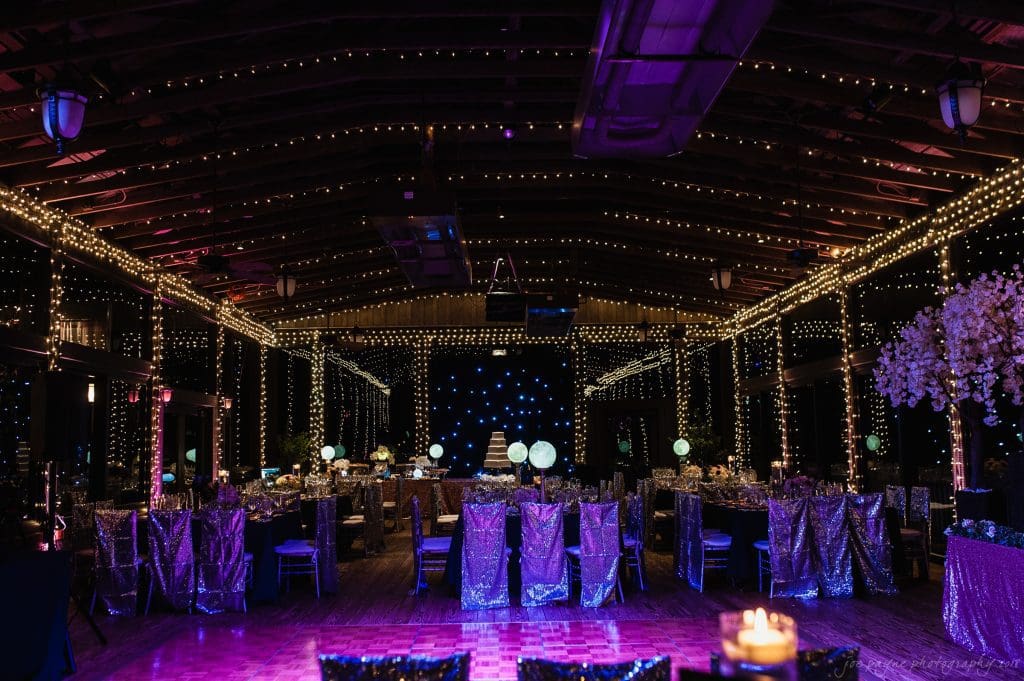 biltmore wedding photographer - anneké & andrew A large, dimly lit event hall is decorated for a formal gathering. Tables are draped with shimmering purple cloths, surrounded by chairs. The room is adorned with twinkling string lights and floral arrangements, creating an elegant and festive atmosphere.